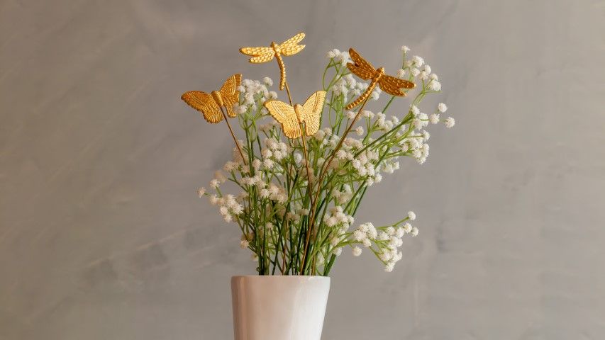 A white ceramic vase filled with delicate white baby’s breath flowers, accented with golden metal decorative sticks shaped like butterflies and dragonflies, against a neutral background.