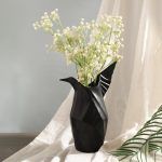 A matte black ceramic vase inspired by origami, designed in the shape of a bird with elegant angular folds. Styled with delicate white flowers, the vase is set against a soft draped fabric, creating a sophisticated and artistic display. Brand: Glimpse Homes.