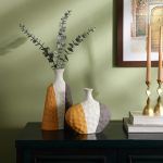 A set of two ceramic vases with a textured surface, featuring a modern color-block design in grey, mustard, and white. The vases have a unique asymmetrical shape and are styled with eucalyptus stems on a dark wooden console against a green wall. Brand: Glimpse Homes.