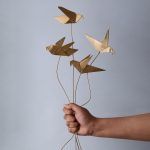 A hand holding a set of 4 golden origami bird metal sticks against a neutral background, showcasing the delicate craftsmanship and bendable stems.