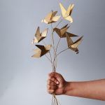 A hand holding a set of 6 golden origami bird metal sticks against a neutral background, showcasing the delicate craftsmanship and bendable stems.