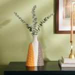 A ceramic vase with a textured surface, featuring a modern color-block design in grey, mustard, and white. The vase has a unique asymmetrical shape and is styled with eucalyptus stems on a dark wooden console against a green wall. Brand: Glimpse Homes.