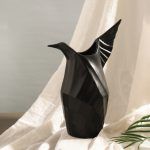 A matte black ceramic vase designed in the shape of an origami-inspired bird, featuring elegant angular folds and a sculptural form. Set against soft draped fabric, the vase exudes a modern and artistic appeal. Brand: Glimpse Homes.