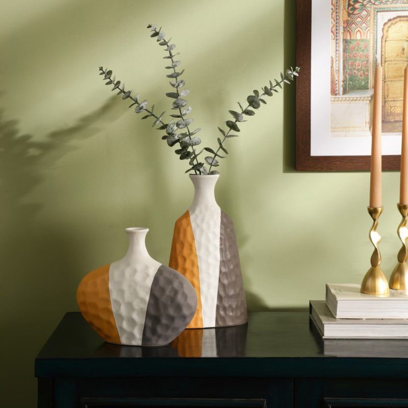 A set of two ceramic vases with a textured surface, featuring a modern color-block design in grey, mustard, and white. The vases have a unique asymmetrical shape and are styled with eucalyptus stems on a dark wooden console against a green wall. Brand: Glimpse Homes.