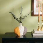 A ceramic vase with a textured surface, featuring a modern color-block design in grey, mustard, and white. The vase has a unique asymmetrical shape and is styled with eucalyptus stems on a dark wooden console against a green wall. Brand: Glimpse Homes.