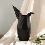 A matte black ceramic vase designed in the shape of an origami-inspired bird, featuring elegant angular folds and a sculptural form. Set against soft draped fabric, the vase exudes a modern and artistic appeal. Brand: Glimpse Homes.