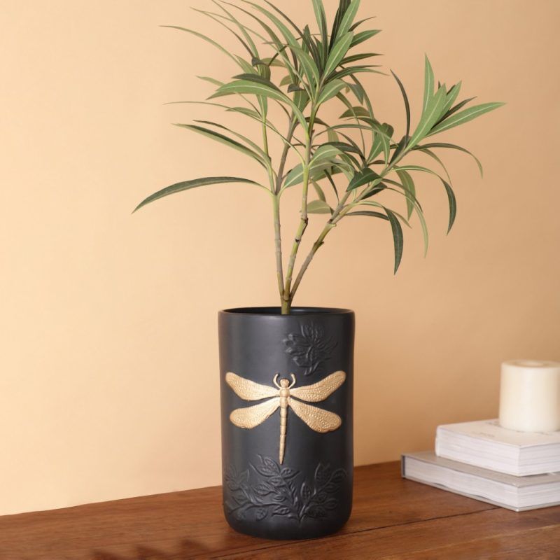 A black matte ceramic vase featuring an embossed floral design and a striking gold dragonfly motif. Styled with fresh green foliage, the vase is placed on a wooden surface against a warm beige background. Brand: Glimpse Homes.