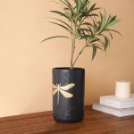 A black matte ceramic vase featuring an embossed floral design and a striking gold dragonfly motif. Styled with fresh green foliage, the vase is placed on a wooden surface against a warm beige background. Brand: Glimpse Homes.