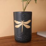 A black matte ceramic vase featuring an embossed floral design and a striking gold dragonfly motif. Styled with fresh green foliage, the vase is placed on a wooden surface against a warm beige background. Brand: Glimpse Homes.