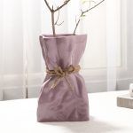 A glossy lilac/lavender ceramic vase with a crumpled paper bag-inspired design, tied with a rustic jute rope for added charm. Styled with delicate dried branches, the vase is placed on a soft white surface near books and a candle, creating an elegant and cozy aesthetic. Brand: Glimpse Homes.