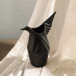 A matte black ceramic vase designed in the shape of an origami-inspired bird, featuring elegant angular folds and a sculptural form. Set against soft draped fabric, the vase exudes a modern and artistic appeal. Brand: Glimpse Homes.