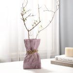 A glossy lilac/lavender ceramic vase with a crumpled paper bag-inspired design, tied with a rustic jute rope for added charm. Styled with delicate dried branches, the vase is placed on a soft white surface near books and a candle, creating an elegant and cozy aesthetic. Brand: Glimpse Homes.