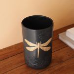 A black matte ceramic vase with an embossed floral design and a prominent gold dragonfly motif. Placed on a wooden surface near books, the vase exudes an elegant and artistic appeal. Brand: Glimpse Homes.