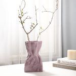 A glossy lilac/lavender ceramic vase with a crumpled paper bag-inspired design, styled with delicate dried branches. Placed on a soft white surface near books and a candle, the vase adds an elegant and modern touch to the decor. Brand: Glimpse Homes.