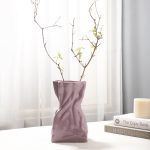 A glossy lilac/lavender ceramic vase with a crumpled paper bag-inspired design, styled with delicate dried branches. Placed on a soft white surface near books and a candle, the vase adds an elegant and modern touch to the decor. Brand: Glimpse Homes.