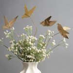 Golden origami bird metal sticks arranged in a white sculptural vase with baby’s breath flowers, creating a modern, Pinterest-worthy home décor setup.