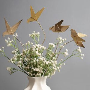 Golden origami bird metal sticks arranged in a white sculptural vase with baby’s breath flowers, creating a modern, Pinterest-worthy home décor setup.