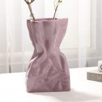 A glossy lilac/lavender ceramic vase with a crumpled paper bag-inspired design, styled with delicate dried branches. Placed on a soft white surface near books and a candle, the vase adds an elegant and modern touch to the decor. Brand: Glimpse Homes.