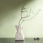 A white ceramic vase with a crumpled paper bag-inspired design, placed on a dark surface against a soft green wall. The vase is styled with a minimalist dried branch, enhancing its organic and artistic aesthetic. Brand: Glimpse Homes.