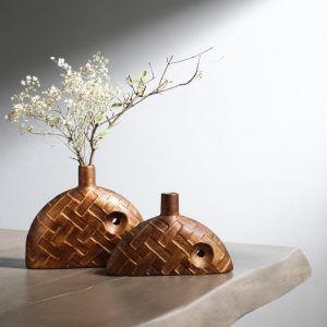 A set of two brown ceramic bud vases with a woven texture design, displayed on a neutral-toned table. One vase is slightly larger than the other, with a small circular cutout detail. The larger vase includes dried floral stems. Brand: Glimpse Homes.