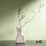 A white ceramic vase with a crumpled paper bag-inspired design, placed on a dark surface against a soft green wall. The vase is styled with a minimalist dried branch, enhancing its organic and artistic aesthetic. Brand: Glimpse Homes.