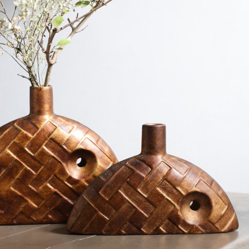 A set of two brown ceramic bud vases with a woven texture design, displayed on a neutral-toned table. One vase is slightly larger than the other, with a small circular cutout detail. The larger vase includes dried floral stems. Brand: Glimpse Homes.