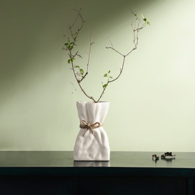 A white ceramic vase inspired by a crumpled paper bag, tied with a rustic jute rope for added charm. Styled with a delicate dried branch, the vase sits on a dark surface against a soft green background, exuding a minimal and organic aesthetic. Brand: Glimpse Homes.