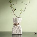 A white ceramic vase inspired by a crumpled paper bag, tied with a rustic jute rope for added charm. Styled with a delicate dried branch, the vase sits on a dark surface against a soft green background, exuding a minimal and organic aesthetic. Brand: Glimpse Homes.