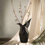 A matte black ceramic vase inspired by origami, designed in the shape of a bird with sculptural folds and a raised wing. Styled with delicate dried branches, the vase is set against soft draped fabric, creating a refined and artistic display. Brand: Glimpse Homes.