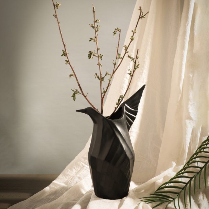 A matte black ceramic vase inspired by origami, designed in the shape of a bird with sculptural folds and a raised wing. Styled with delicate dried branches, the vase is set against soft draped fabric, creating a refined and artistic display. Brand: Glimpse Homes.