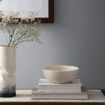 A white ceramic bowl placed on stacked books, styled with a ceramic vase and framed artwork for a contemporary decor setting.