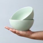 A pair of stacked matte-finish sage green ceramic thukpa soup bowls balanced on an open hand against a soft blue background.