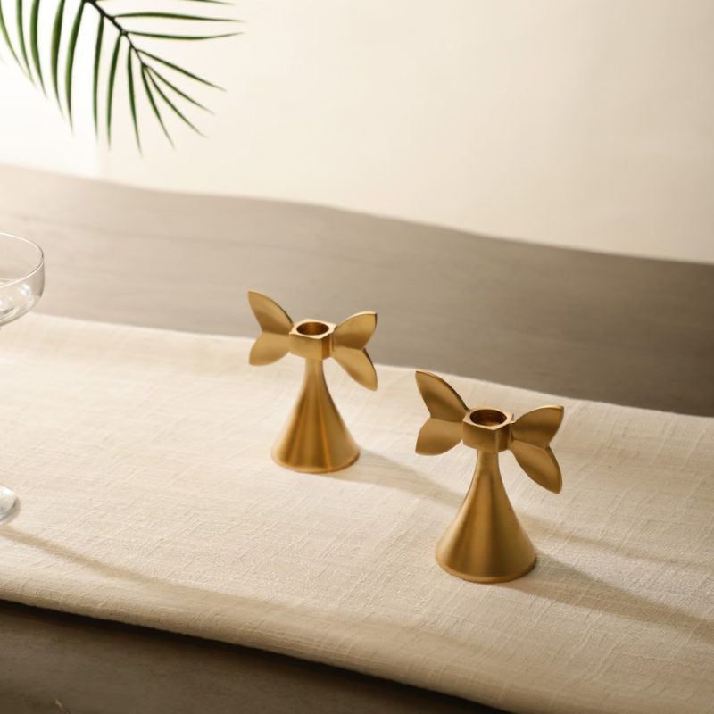 A pair of gold-finished metal candle holders with an elegant bow-shaped design, placed on a neutral linen table runner. The warm lighting highlights their smooth metallic texture, creating a sophisticated and timeless ambiance.