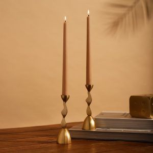 A pair of gold metal candle holders with fluid, sculptural stems, supporting soft beige taper candles. The warm-toned background creates an elegant and timeless aesthetic.