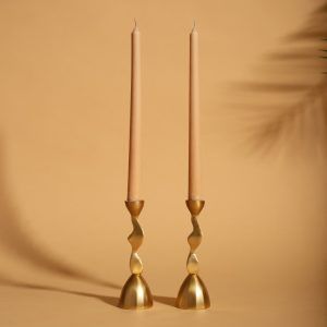 Two contemporary gold metal candle holders with an artistic wavy design, each holding a beige taper candle. A minimalist warm backdrop highlights their modern elegance.