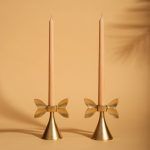 Two gold metal candle holders with a bow-inspired design, each holding a slender beige taper candle. Set against a warm-toned background, the holders exude a charming and modern aesthetic, perfect for refined table settings.