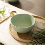 Matte-finish sage green ceramic bowl placed on a neutral-toned tray with a napkin and a gold spoon beside it.