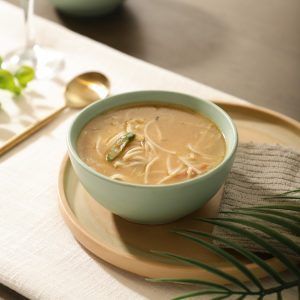 Sage green ceramic bowl filled with steaming Thukpa, served on a neutral-toned tray with a napkin and gold spoon.