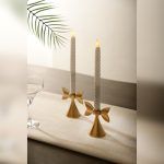 A pair of Golden Bow Candle Holders, crafted from lacquered aluminium, elegantly displayed with white twisted LED taper candles on a linen table runner. The soft lighting enhances their refined charm.