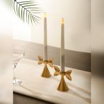 Two Golden Bow Candle Holders with a delicate bow-shaped design, holding smooth white LED taper candles. Placed on a neutral table setting, they create a modern yet timeless aesthetic.