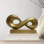 Infinity loop metal accent piece in antique gold finish placed on a metal base, displayed on a stack of books with neutral background.