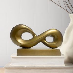Infinity loop metal accent piece in antique gold finish placed on a metal base, displayed on a stack of books with neutral background.
