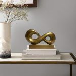 Infinity loop metal knot in antique gold finish placed on a stack of books on a side table, next to a ceramic vase with white flowers and a framed artwork on the wall.