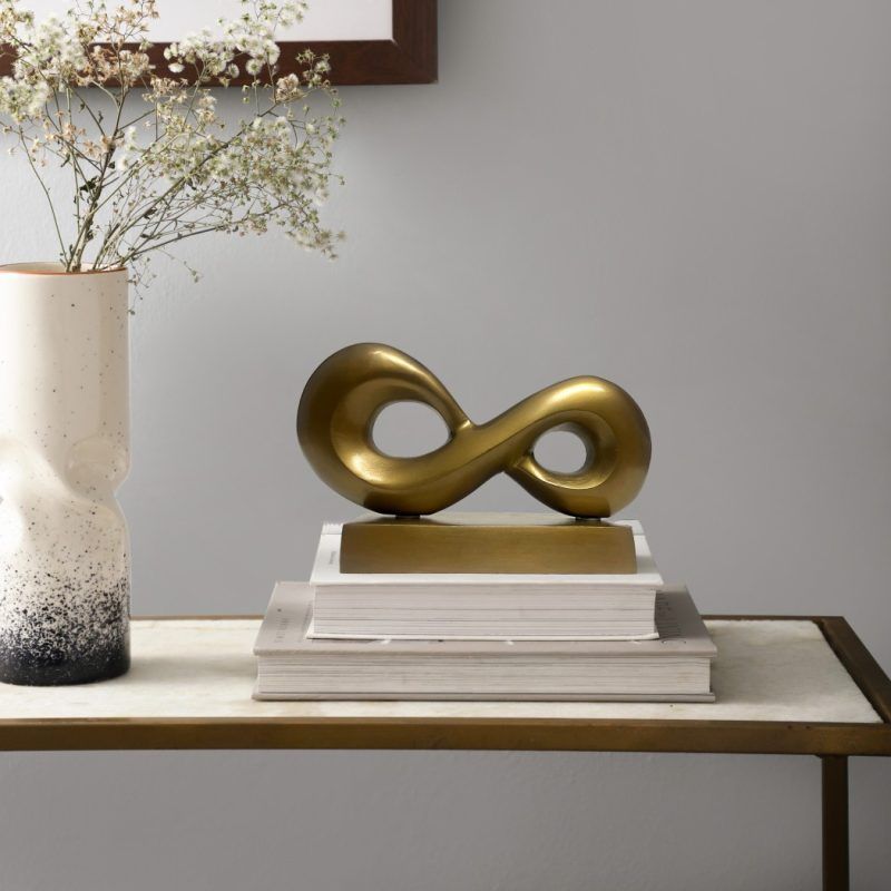 Infinity loop metal knot in antique gold finish placed on a stack of books on a side table, next to a ceramic vase with white flowers and a framed artwork on the wall.