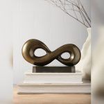 Infinity loop metal accent piece in antique brass finish placed on a matte black metal base, displayed on a wooden surface with neutral background.