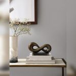 Infinity loop metal knot placed on a stack of books on a side table, next to a ceramic vase with white flowers and a framed artwork on the wall.
