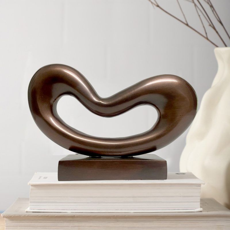 A modern Wave Form Metal Sculpture in a copper finish sits on a stack of white books. The sculpture features a smooth, avant-garde wave design with a central cutout, set against a minimalist background with a white vase and dried branches.