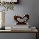 A Wave Form Metal Sculpture in copper, displayed on a wooden base atop a stack of white books on a light-colored table. The sculpture's sleek, modern wave design is shown alongside a white speckled vase with dried branches, a framed picture, and a minimalist background.