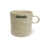 A beige ceramic mug with a textured surface and a handle, displaying the text "DREAMER." in black capital letters with a period, placed against a white background.
