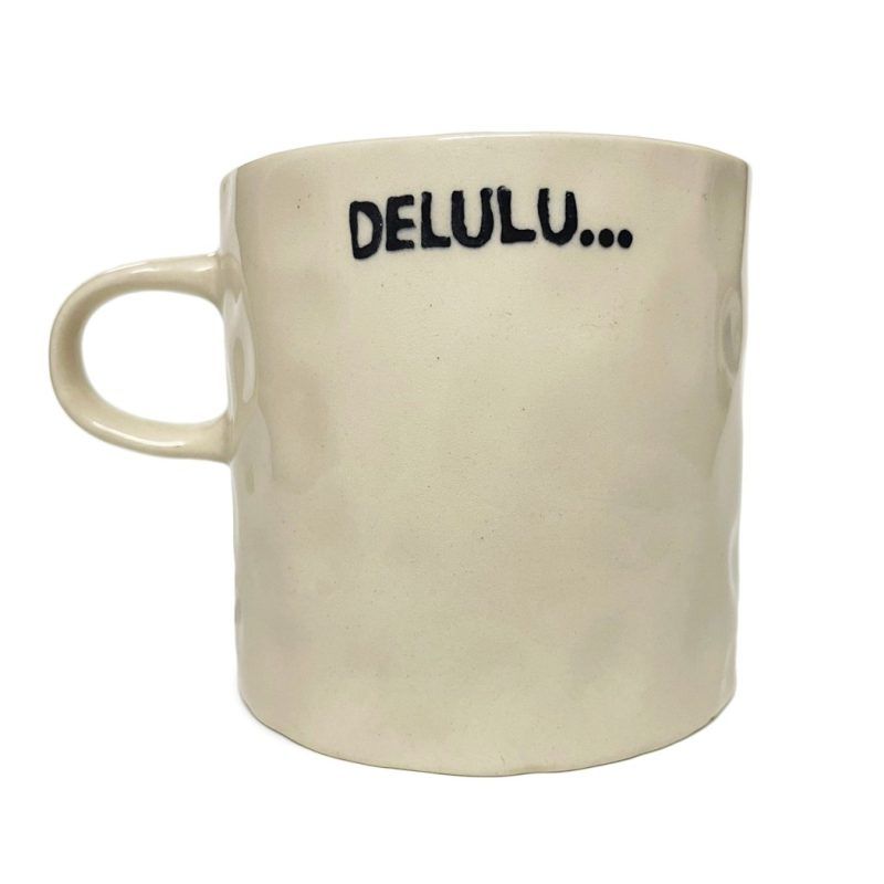 white background image of an off-white, textured ceramic mug. The mug has the word “DELULU…” printed in bold black letters.